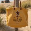 sac plage xl jaune Grand sac cabas Sel de Mer en toile recyclée jaune safran suspendu à un poteau en bois sur une plage landaise. Détails en cuir et impression vintage sur le devant